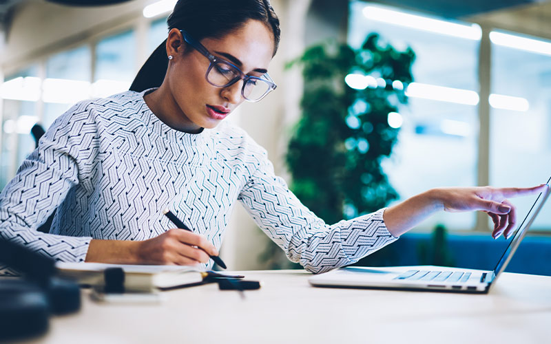 Woman making notes while looking at laptop