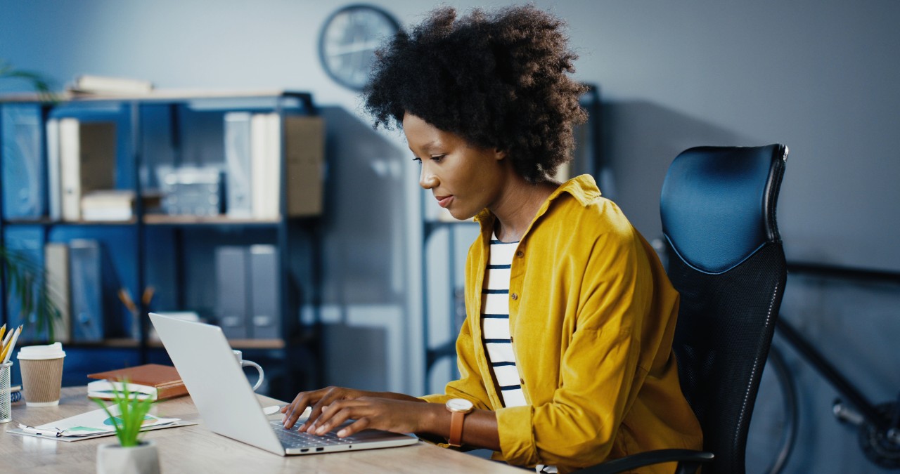 Beautiful joyful curly female worker typing on laptop and searching internet in cabinet. Pretty concentrated African American businesswoman working on computer while sitting at office. Job concept