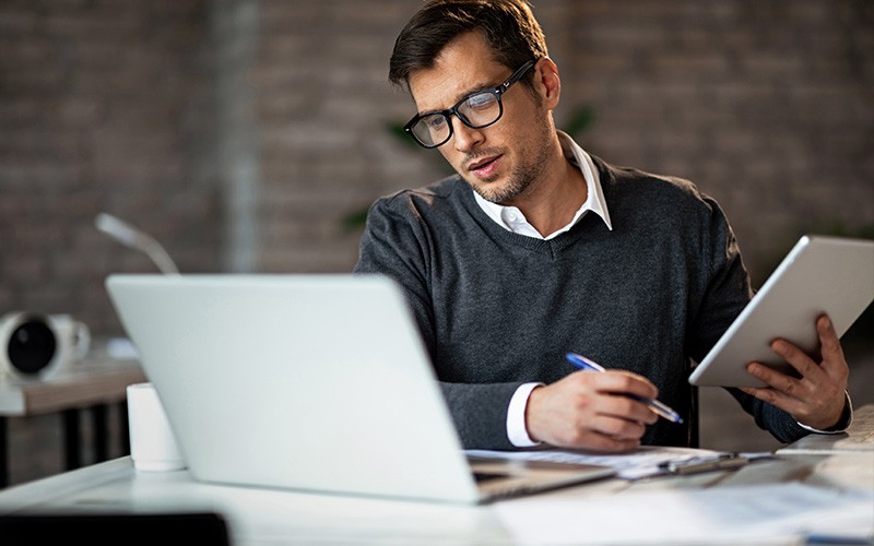Man holding a tablet while looking at a laptop